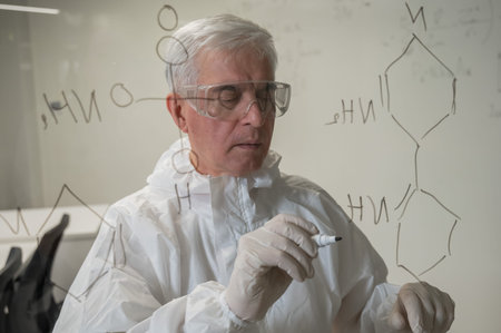 An elderly Caucasian male chemist in a protective suit writes on glass
