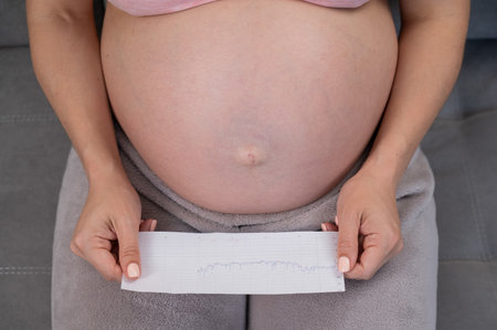 A pregnant woman looks at the results of cardiotocography while sitting on the couch. Close up of the bellyの写真素材