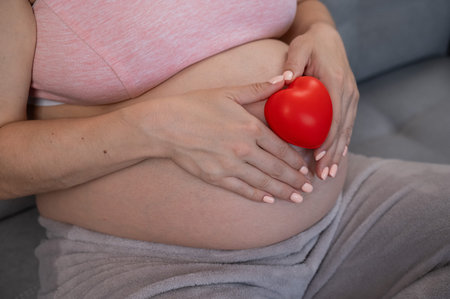 Caucasian pregnant woman holding heart on belly background. Third trimester