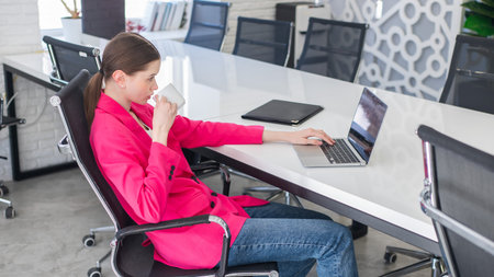 Caucasian business woman in fuchsia jacket drinking coffee at her deskの写真素材