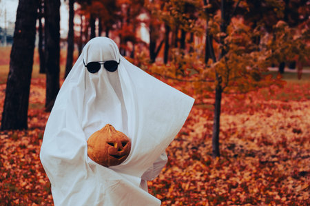 Woman in white sheet holding jack olantern against autumn forest background. Halloween costume