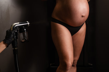 Faceless pregnant woman getting an instant tan. The master applies bronzer with a sprayの写真素材