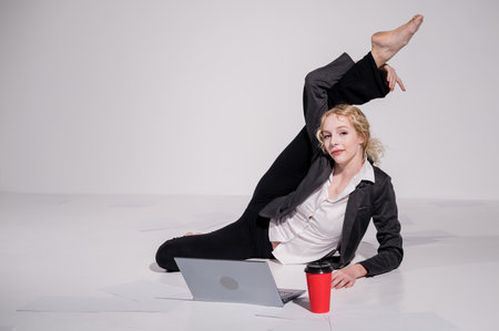 Barefoot ballerina dressed in a business suit poses for a laptop and drinks coffee. Flexible woman works at the computer.の写真素材