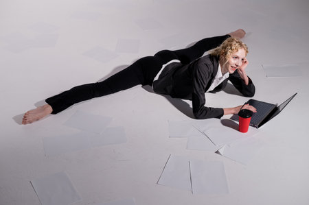 A ballerina dressed in a business suit sits in a middle twine, works at a laptop and drinks coffee. Flexible woman works at the computer.の写真素材