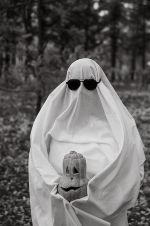 Woman in white sheet holding jack olantern against autumn forest background. Halloween costume. Black and white photoの写真素材