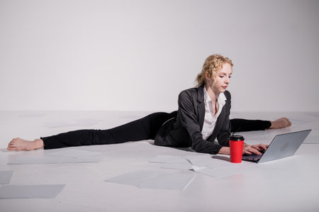 A ballerina dressed in a business suit sits in a middle twine, works at a laptop and drinks coffee. Flexible woman works at the computer.の写真素材