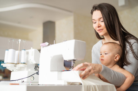 A young woman sews in the kitchen and holds a small child. Mom teaches her little son to sew on a sewing machine. Self-isolation homework.の写真素材