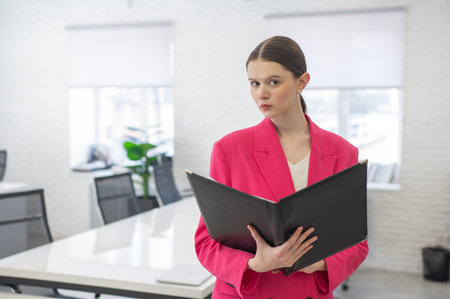 Smiling business woman in pink jacket reading documents in officeの写真素材