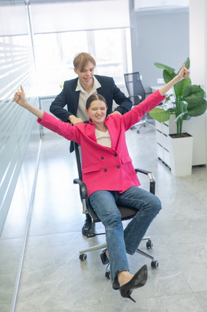 Coworkers fooling around in the office. A guy pushes a young woman around on a chairの写真素材