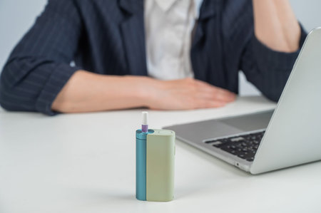 Business woman smoking an electronic cigarette while working on a laptop. Tobacco heating system in...