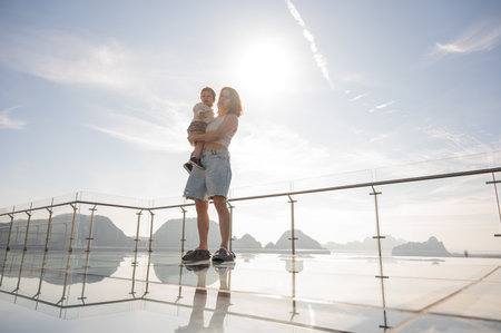 Caucasian woman holds her little son in her arms while standing on a glass bridge in the mountainsの写真素材