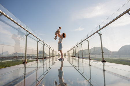 Caucasian woman holds her little son in her arms while standing on a glass bridge in the mountainsの写真素材