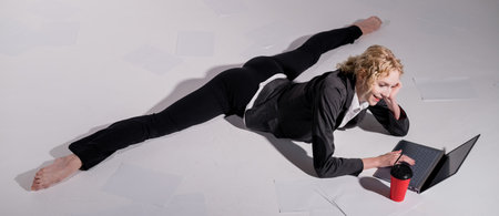 A ballerina dressed in a business suit sits in a middle twine, works at a laptop and drinks coffee. Flexible woman works at the computer.の写真素材