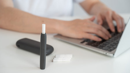 A woman smokes an electronic cigarette while working on a laptop. Tobacco heating system