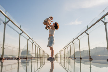 Caucasian woman holds her little son in her arms while standing on a glass bridge in the mountainsの写真素材