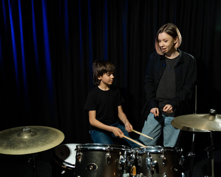 Young caucasian woman teaches a boy to play the drums in the studio on a black background. Music school student.の写真素材