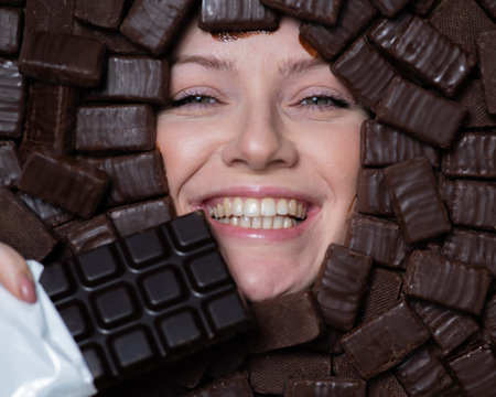 The face of a caucasian woman surrounded by chocolates. The girl eats a bar of chocolate