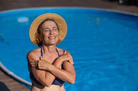 An elderly woman in a straw hat is sunbathing by the pool. Retiree on vacation