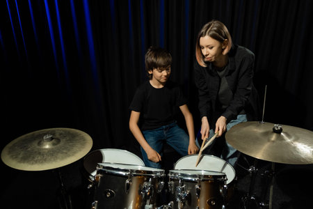 Young caucasian woman teaches a boy to play the drums in the studio on a black background. Music...