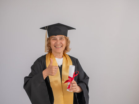Happy senior woman in graduation gown holding diploma on white backgroundの写真素材