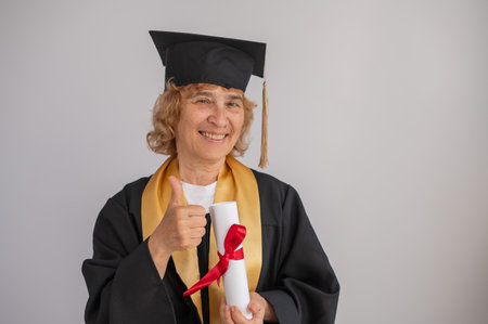 Happy senior woman in graduation gown holding diploma on white backgroundの写真素材