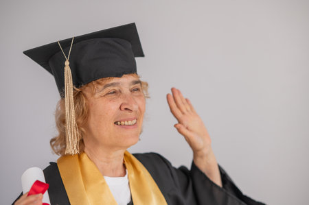 Happy senior woman in graduation gown holding diploma on white backgroundの写真素材