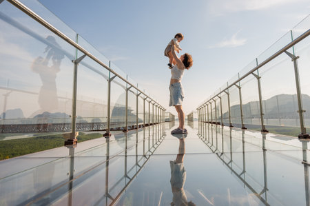 Woman holds her little son in her arms while standing on a glass bridge in the mountainsの写真素材