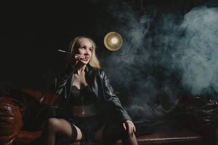 A woman smokes from a mouthpiece while sitting on a leather sofa in her underwearの写真素材