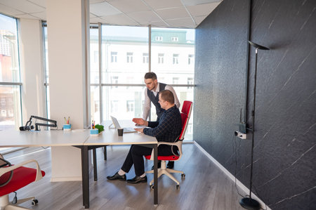 Caucasian man standing at colleagues desk discussing work