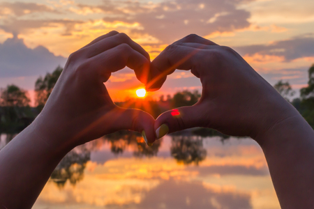 Hands in the form of heart on the background of the sun at sunsetの写真素材