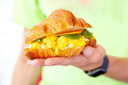 Hand holding a breakfast brioche croissant sandwich with scrambled egg filling cheese and rocket leaf.の写真素材