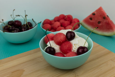 Vanilla ice cream with watermelon balls and cherries in a light turquoise bowl on a bamboo boardの写真素材