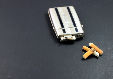 A vintage cigarette lighter/case next to a pile of cigarette butts on an old black tableの写真素材