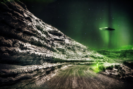 Alien on a lonely dirt road at night with a UFO flying off in the background.の写真素材