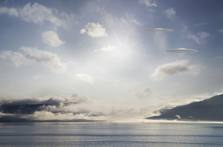 Two UFOs hovering over a coastal scene with clouds and sun.の写真素材