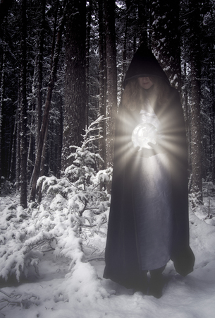 Woman in a long hooded cape in a winter forest holding a glowing crystal ball.の写真素材