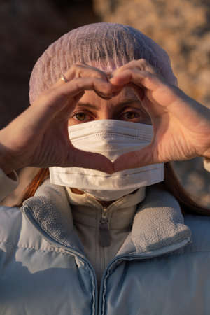 A girl in a medical mask and hat, on a sunny day, holds her fingers with a heart, pandemicの写真素材