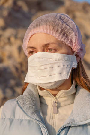 Redhead girl in a medical mask and hat in bright clothes on the street on a sunny day, pandemicの写真素材