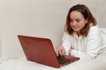 A young girl lies at home on the sofa with a computer and communicates via Skype with friends, making hand gestures. covid-19 epidemic concept. Quarantine stay at home.の写真素材