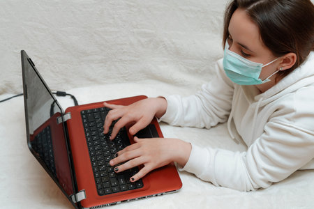 A girl in a mask in a white hoodie is holding a laptop and typing on the computer. free space. We...