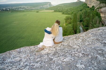 A man and a woman sit with their backs embraced on a high rock and look at the landscape. Green field with a river on the right and mountains on the horizon. The concept of romance, travel, adventureの写真素材