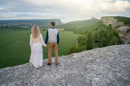 A man and a woman are standing with their backs on a high rock holding hands and looking at the...