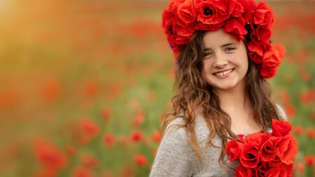Portrait of a beautiful teenager girl in a wreath of poppy flowers looking smiling, on a green field with a bouquet of poppies in a summer sunset.の写真素材