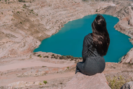 A brunette girl with loose hair in a gray dress sits on the background of an azure lake in the form...