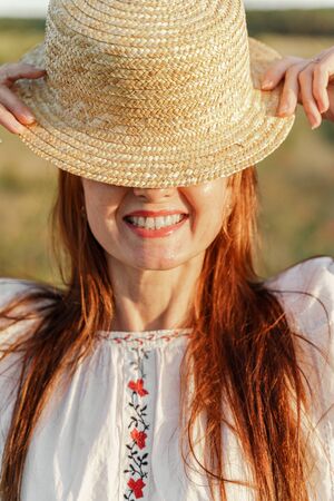 A red-haired smiling woman holds a hat in her hands, only the bottom of her face with a smile is visible. On a green field.の写真素材