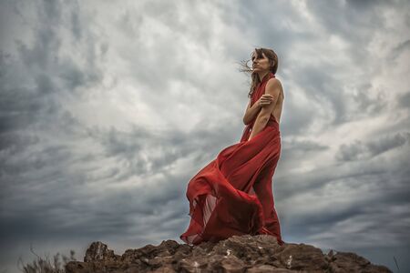 Side view a beautiful sensual woman in a red long dress posing on a rock high above the sea during sunset. Dark clouds all over the skyの写真素材