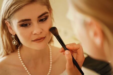 Make-up artist makes professional makeup of a young woman, a bride. The bride in a white dress, a pearl necklace and earrings. Make-up artist applies powderの写真素材