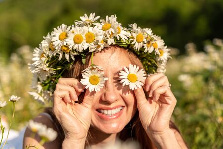 A funny young woman in a wreath of daisies is laughing and holding daisies in front of her eyes. On a large field of daisies against the background of the forest.の写真素材