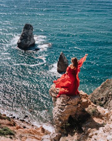 A girl with loose hair in a red dress waved her skirt on the yellow rocks overlooking the sea. Sunny path on the sea from the rising sunの写真素材