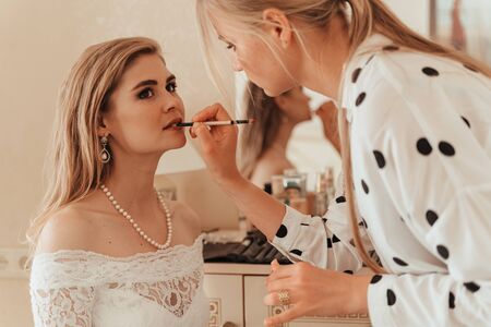Make-up artist makes professional makeup for a young woman, a bride. The bride in a white dress, a pearl necklace and earrings. Make-up artist paints the bride's lipsの写真素材